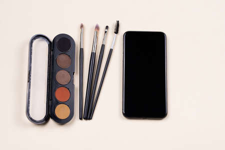 Online course of make up study. Palette of eyeshadows, cosmetics brushes and smartphone empty screen on light beige background. Beauty and fashion webinarの写真素材