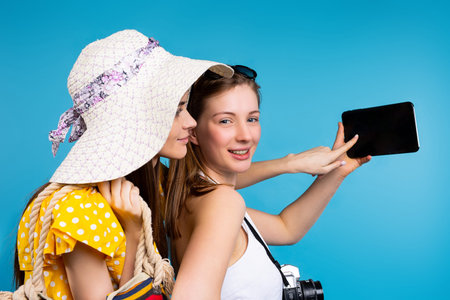A photo from the side of two girlfriends in summer clothes on a blue background are holding a tablet...