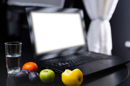 Glass of water and fresh fruits near empty white mock up laptop screen. Healthy snack and working in the office. Bitten apple, orange and plums on table. Healthy online blogging. High quality photoの写真素材