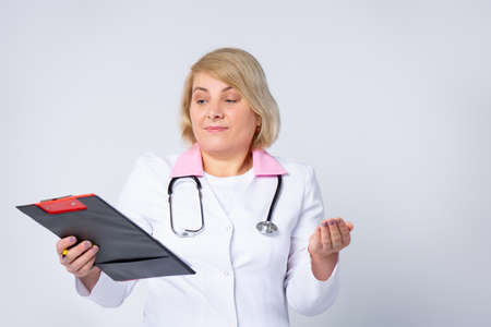 Senior handsome doctor, woman with blonde hair, wearing medical uniform, looking on sheet folder with prescription. Confused expression with arms and hands raised. Doubt concept. High quality photoの写真素材