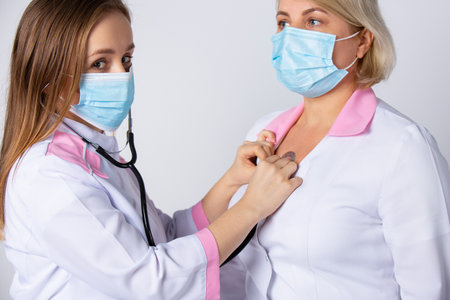 Young doctor using stethoscope listen senior doctor heart during examination in hospital. Medical control for infections, disease on gray background. Suffering medical staff. High quality photoの写真素材