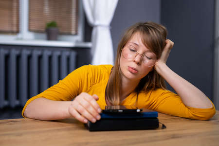 Fatigued blonde girl with glasses sitting at home with electronic book on digital tablet, doing homework, looking down. Bored student got tired of studying, missing her friends. High quality photoの写真素材