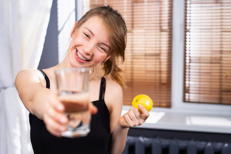 A blurry photo where a girl with a smile holds out a glass of clean water to the camera and holds a lemon in her hand. Benefits of water for fasting. Proper nutrition. High quality photoの写真素材