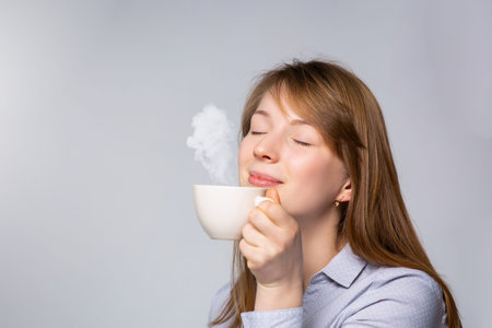 Smiling woman holding steaming coffee cup against gray background. Hot tea time, home in the...