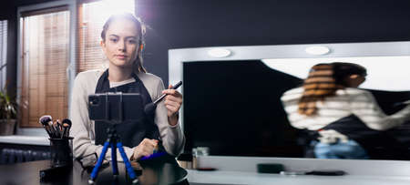 Banner,long format. A woman beauty blogger shoots her daily makeup tutorial on camera. A female video blogger applies powder with a brush in front of a large professional mirror. High quality photoの写真素材
