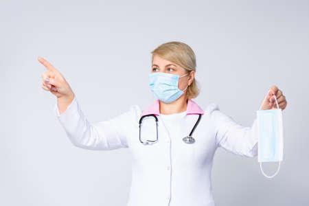 Middle-aged female doctor in respirator mask with raised forefinger. Portrait of confident doctor with stethoscope holding protective mask isolated on white background. High quality photoの写真素材
