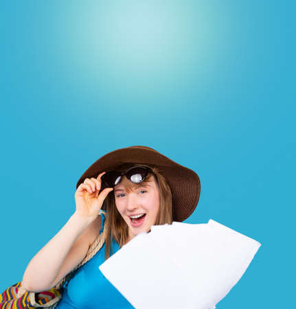 Banner. Portrait of a happy girl in a hat who takes off her glasses and shows empty tickets to the camera. Tourism concept on blue background with blank top space. High quality photoの写真素材