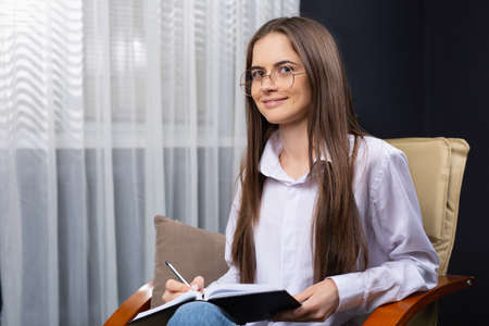 Brunette woman in white shirt sits on leather chair in home office, holds pencil, black notepad. Millennial caucasian girl taking notes in notebook. Woman drawing up business plan. High quality photoの写真素材