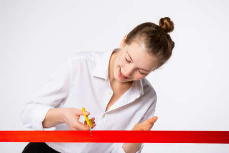 Amazing young businesswoman cutting red ribbon with a pair of scissors of illuminating color. Caucasian confident woman launch new project, store. Grand opening on gray background. High quality photoの写真素材