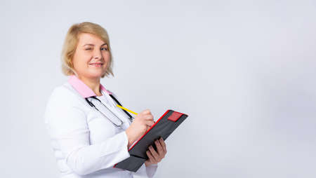 Banner. Portrait, an attractive senior female doctor, white coat with sthetoscope, writes analysis, black clipboard, illuminating pencil. Gray background with plenty space for ad. High quality photoの写真素材