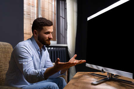 Outraged man at home sitting on the couch in front of a large TV screen. High quality photoの写真素材