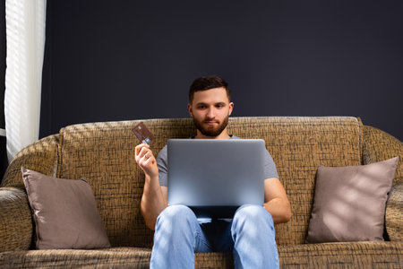 A young man in home clothes is sitting at home on a comfortable sofa with a laptop. Works and holds a bank card. Work online. Transfer of earned money. High quality photoの写真素材