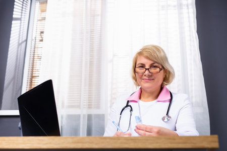 Portrait of a senior woman doctor in a medical gown and glasses sitting at the table, he uses a...
