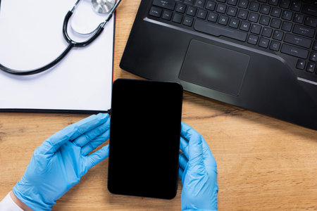 Hands of a paramedic in latex gloves holding a tablet. On the table a laptop, paper and a...