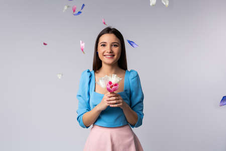 A beautiful brunette holds tulips in her hands and smiles at the camera. Gray background and falling colorful petals. Side advertising space.の写真素材