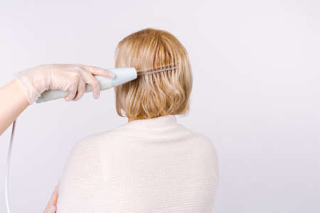 Photo back where the beauticians hand makes hair procedures for a blonde woman with the help of darsonval. White background and side space.の写真素材