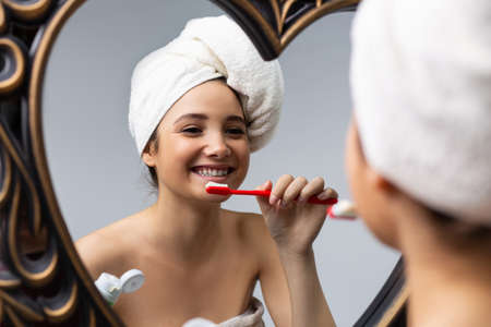 Morning oral care. A girl with a towel on her head brushes her teeth in front of a mirror. Reflection. Close-up.の写真素材