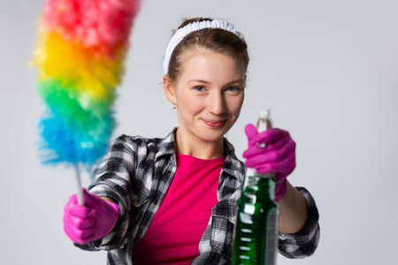 Portrait of happy young housewife doing the household chores holding a dust brush and disinfectant...