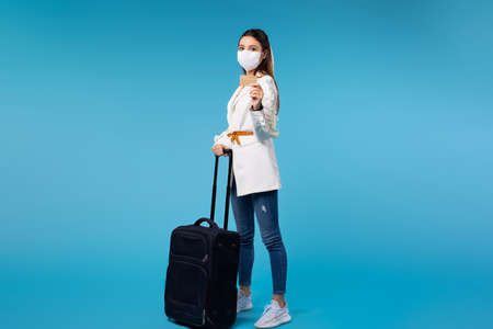 Young caucasian woman in medical mask with suitcase looking to the camera while standing near modern gates on railroad station, showing bank card. Studio photo on blue backgroundの写真素材