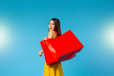 Caucasian woman holding red shopping bags. Excited female shopaholic, summer holiday sales promotion advertisement marketing on blue isolated background with blank copyspaceの写真素材