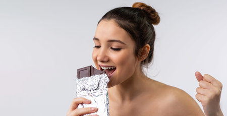 Banner- long format, grey background. Young smiling caucasian woman eating bar of dark chocolate....