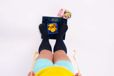 Obese woman standing on scales to check body weight along with a plate of cookies, overweight problem. Weakness for sweets concept.の写真素材