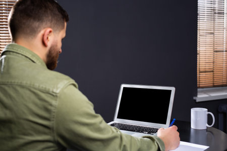 A photo from the back of a man sitting at a writing desk. Fills in documents and works with a laptop...