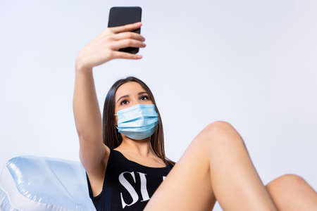 A brunette girl in a swimsuit and a medical mask on an inflatable mattress makes a selfie on a smartphone. Rest during a pandemic on a white background.の写真素材