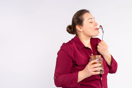 Caucasian chubby woman holding a glass jar and happily eating shekolade pasta with a large spoon....