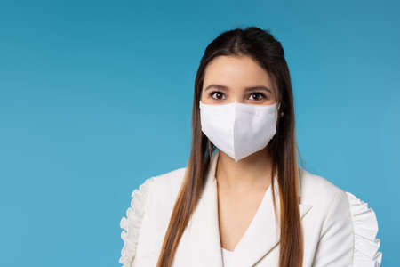 Portrait of a young serious lady in a white protective mask on a blue background looks at the camera. Office work and business during a pandemic.の写真素材