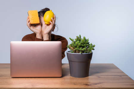 A brunette sitting at a wooden table covers her face with a sponge and lemon. The concept of ecological home remedies. Gray background and side space.の写真素材
