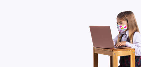 Banner,long format. Caucasian schoolgirl in uniform and a medical mask sits at a table with a laptop. The concept of training. Preventing the spread of the virus.の写真素材