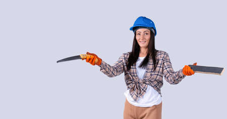 Breaking gender stereotypes. Confident woman in a blue safety helmet holding a board and a hand saw. Studio gray background.の写真素材