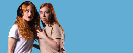 Banner. The pastime of girlfriends. Red-haired girls listen to music on big headphones isolated on studio blue background.の写真素材
