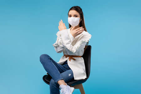 A young girl in a white blouse and a medical mask sits in an office chair and on a blue background shows stop, the end of the spread of the virus. Work and pandemic.の写真素材
