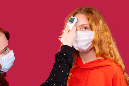 An enlarged photo of two girls in a medical mask measuring the temperature with a non-contact thermometer at the forehead. Red background. Protective measures during a pandemic.の写真素材