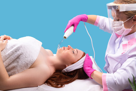 A woman beautician in a white coat, in a medical mask and a protective screen makes dorsanvalization of the skin of the face for a young client wrapped in a towel.の写真素材