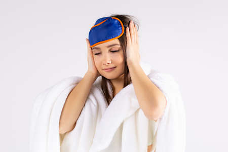 Young beautiful woman with sleep mask having headache, injury, fatigue on grey studio portrait. Sad girl putting hands on head, isolated. Concept of problems and headache.の写真素材