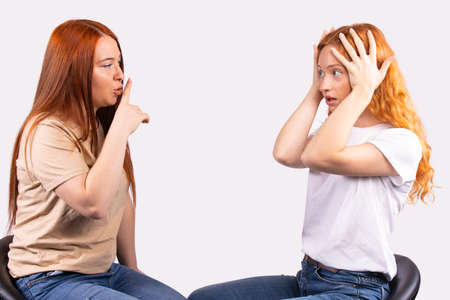 Young pretty red haired girl making silence gesture, hush with finger on lips to her frightened, scared friend, asking to be quiet. Silence or secret concept on gray background, free space for textの写真素材