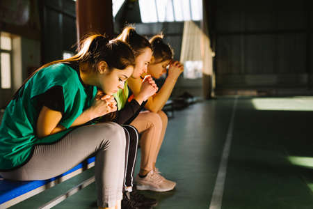 Players praying for success waiting final goal. Sportswomen prays with team for winning indoor gym arena court sitting on the spare bench.の写真素材
