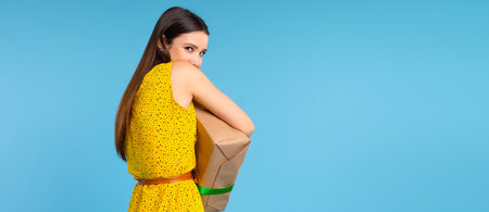 Banner, long format. A photo from the back of a girl in a yellow dress who is hiding a large gift box in front of her. Blue background and advertising space. High quality photoの写真素材