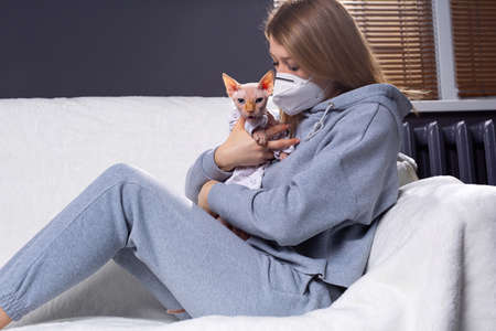 Tired and sad woman wearing a protective face mask cuddles, plays with her furious and mad cat at home. Female owner suffer of coronavirus pandemic covid-19. High quality photoの写真素材