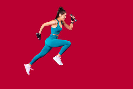 Athletic young girl in sports uniform and white snickers makes lunges with dumbbells in her hands over the red background. High quality photoの写真素材