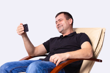 Adult man with hearing aid sitting in a chair holding a telephone in his hand and talking with somebody laughing. High quality photoの写真素材