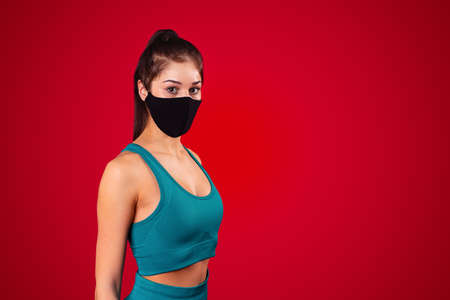 A sportive young woman is posing in a black multifunctional mask dressed in sports uniform over red background. High quality photoの写真素材