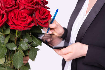 A woman is holding the tablet and is ready to sign with the blue pen for delivery of red roses by courier. High quality photoの写真素材