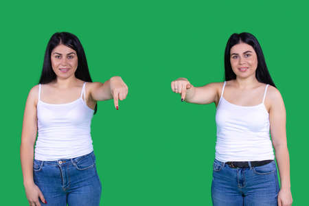 Two amazing beautiful girls are standing on distance and showing down with their index finger over a green background.の写真素材