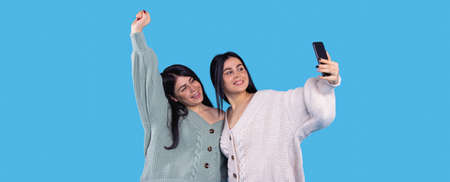 Cheerful twin girls making selfie isolated on blue background one of them raised her right hand up and smiling.の写真素材
