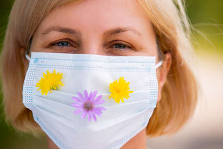 Close up photo of a senior blonde woman looking right in front of the camera wearing medical mask with meadow flowers on the mask, takes care of her health outdoors.の写真素材