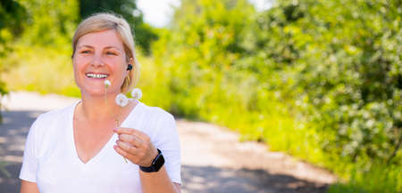 Close up photo of a happy blonde woman against a background of green foliage in nature, holding the dandelion flower she found after fitness exercises.の写真素材